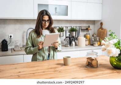 Smiling young woman wearing glasses drinking coffee and using tablet computer while sitting in kitchen. Businesswoman reading emails at morning in her home. Remote work business female - Powered by Shutterstock