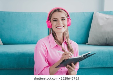 Smiling young woman wearing headphones and writing in notebook while relaxing at home in bright living room - Powered by Shutterstock