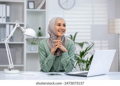 Smiling young woman wearing hijab and working on a laptop at her desk. Modern office setting with a bright, professional atmosphere. - Powered by Shutterstock