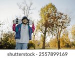 Smiling young woman wearing protective helmet carrying skateboard on shoulders in park with autumnal trees enjoying sunny day