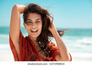 Smiling young woman wearing red lace dress and looking at camera. Beautiful tanned girl enjoying summer vacation at sea. Portrait of stylish carefree woman laughing at the ocean with copy space.