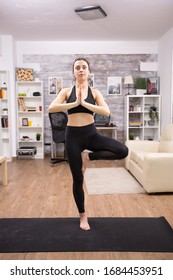 Smiling Young Woman While Doing Tree Yoga Pose Wearing Black Sportwear In Home.