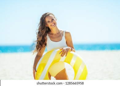 Smiling Young Woman In White Beachwear With Yellow Inflatable Lifebuoy On The Beach. Getting Vitamin D After Long Winter Months. сaucasian Woman With Long Brunette Hair 30 Something Years Old.