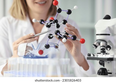Smiling young woman in white lab coat demonstrates model of molecule using set of 3D atoms. Doctor explains chemical properties of elements at lesson