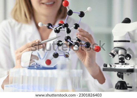 Smiling young woman in white lab coat demonstrates model of molecule using set of 3D atoms. Doctor explains chemical properties of elements at lesson