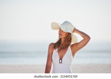 A smiling young woman in a white swimsuit holding her hat and enjoying a day at the beach.A carefree young woman in swimwear thinking and relaxing by the sea. A woman on holiday at the beach