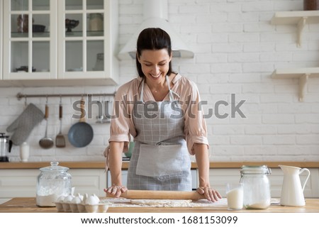 Smiling young woman work with wooden roller pin making sweet pie or pastry dough in kitchen, happy millennial female loving wife cooking in apron preparing family dinner or dessert baking buns