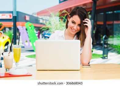 Smiling Young Woman Working on Laptop and speaking on the Smart Phone at Shopping Mall while Drinking Juice