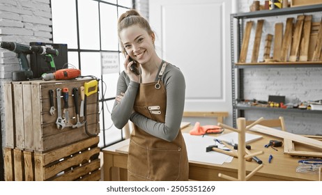 A smiling young woman in a workshop making a phone call while wearing casual clothes and a work apron surrounded by tools. - Powered by Shutterstock