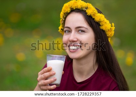 smiling young woman with wreath of dandelion flowers drinking yogurt from glass outdoors on the meadow. Portrait of girl with funny milk mustache