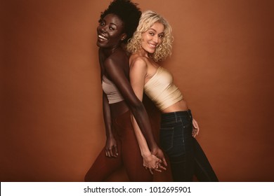 Smiling Young Women Standing Back To Back And Looking At Camera On Brown Background. Diverse Female Models Standing Together And Smiling.