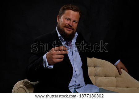 Smilingl solid bearded man dressed in suit sitting on sofa studio portrait on black background