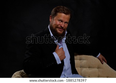 Smilingl solid bearded man dressed in suit sitting on sofa studio portrait on black background