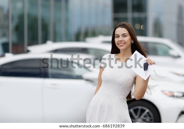 Smilling woman with keys and agreement on\
background of car\
dealership