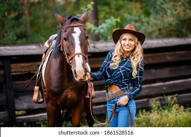 Smilng pretty young cowgirl. A female in a plaid shirt and a cowboy hat standing next to the horse and holds the horse by the bridle.