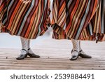 Smiltene, Latvia - October 27, 2024 - Close-up of two women’s legs wearing colorful striped skirts, lace socks, and black shoes, showcasing traditional attire on a wooden platform.