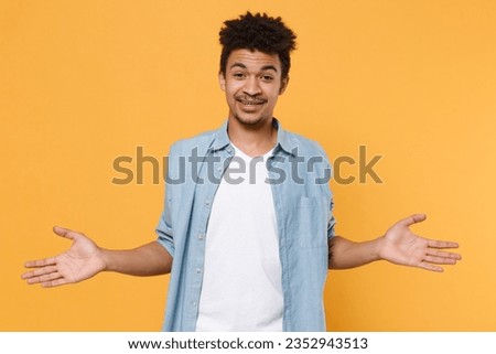 Smirked perplexed young man of African American ethnicity in casual blue shirt posing isolated on yellow wall background studio portrait. People lifestyle concept. Mock up copy space. Spreading hands