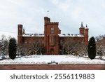 The Smithsonian Castle in Washington, D.C., during winter, surrounded by snow-covered gardens and a serene atmosphere under a cloudy sky.