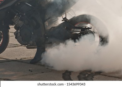 The Smoke Comes Out From Under The Wheels. Motorcycle Wheel Closeup. Smoke Due To Tire Rubbing Against Asphalt. The Rider Prepares To Do The Trick On The Motorcycle. Burned Rubber On The Road.