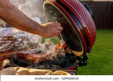 Smoke coming out of red ceramic Barbecue Grill. The man coats pork ribs with barbeque sauce. Picnic in modern homes terrace. - Shutterstock ID 1694266516
