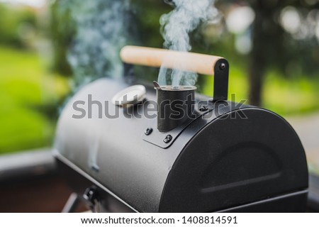 Smoke coming out of a smokestack of a small black smoker grill or barbecue on green background.