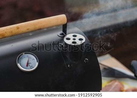 Smoke coming out of a smokestack of a small black smoker grill or barbecue, Visible also temperature gauge and wooden opening handle.