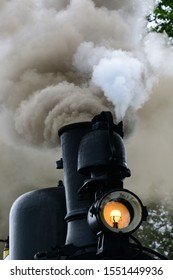 Smoke Stack Of A Narrow-gauge Steam Train With A Lot Of Grey And White Smoke And The Locomotive Front Light