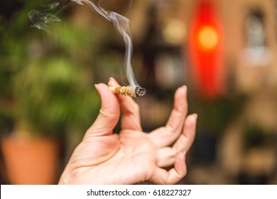Smoke streams from the end of a burning joint in a woman's hand. Joint is covered in oil and kief or THC powder.