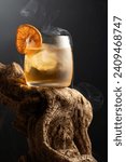 Smoked old fashioned rum cocktail with ice and dried orange slice on a old wooden snag. Copy space.