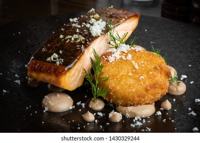 Smoked Salmon Steak With Parmesan Cheese Crumb And Crab Fishcake Presented On A Black Plate With Garnish.