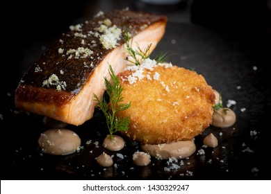 Smoked Salmon Steak With Parmesan Cheese Crumb And Crab Fishcake Presented On A Black Plate With Garnish.