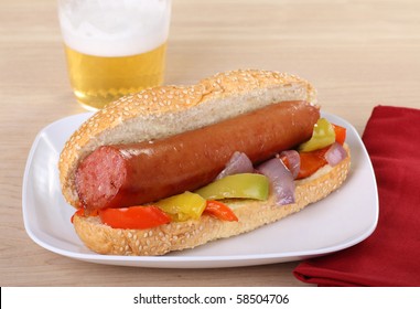 Smoked Sausage On A Sesame Seed Bun With Peppers And Onions And A Beer In The Background