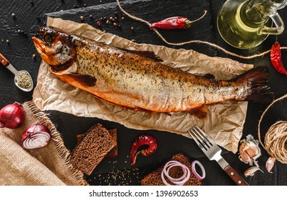 Smoked trout fish, red whole fish with spices and onion on craft paper over dark stone background. Seafood, top view