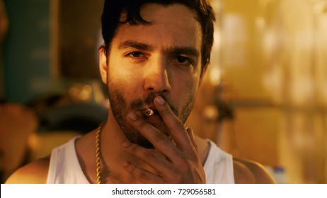 Smoking Brutal Gang Member With Gold Chain Looks Into Camera With Defiance. Underground Drug Laboratory Is In Background.