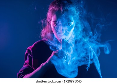 Smoking an electronic cigarette. The girl's face is covered with smoke from vape. Portrait of a female smoker in a cloud of smoke. A woman smokes a VAPE on a dark background.