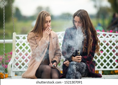 Smoking And Vaping. Young Beautiful White Teeage Girls In Casual Clothes Sit On A Vintage Bench And Smoke And Vape Cigarettes On The Street In The Evening. Electronic Cigarette. Bad Habit.