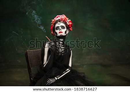 Smoking. Young girl like Santa Muerte Saint death or Sugar skull with bright make-up. Portrait isolated on dark green studio background with copyspace. Celebrating Halloween or Day of the dead.