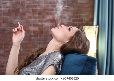 Smoking Young Woman Sitting In Armchair And Blowing Cigarette Smoke In Living Room