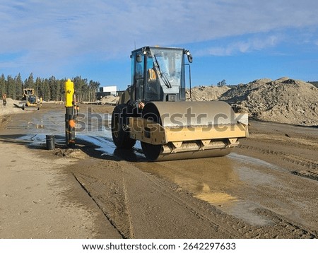 Smooth drum roller compacting wet subgrade near installed fire hydrant at active subdivision site under clear sky.