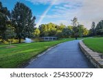 a smooth footpath through the park surrounded by green grass and lush green trees with a lake with a water fountain and a stone bridge, blue sky clouds and a rainbow at Lenox Park in Brookhaven