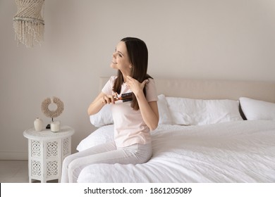 As smooth as silk. Glad young woman sitting on bed at morning taking care of long hair feeling content with its shiny healthy look. Combing with professional circle hairbrush having natural bristles