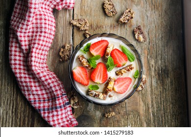 Smoothie Yogurt Bowl With Mint, Strawberries, Chocolate Granola Bar On The Dark Wood Background With A Kitchen Towel, Top Down View