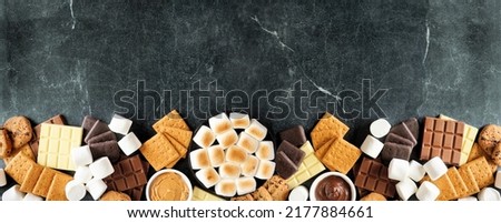 Smores buffet bottom border. Roasted marshmallows, crackers, chocolate and a assortment of ingredients. Top view over a dark stone background.