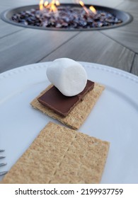 Smores Ingredients On White Plate In Front Of Fire Table.