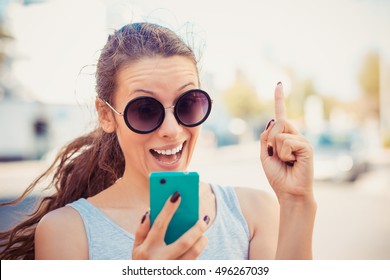 SMS. Closeup Portrait Happy Pleased Young Girl Having Idea Pointing Up Finger Gesture Looking At Phone Seeing Good News Photos Message Disgusting Emotion On Face Isolated Cityscape Outdoor Background