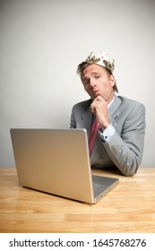 Smug Businessman Wearing A Crown Sitting At His Desk Looking At The Camera