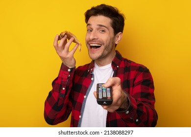 Snack And Entertainment. Happy Guy Holding Slice Of Pizza Watching TV Switching Channels With Remote Control In Hand Posing On Yellow Orange Studio Background, Looking At Camera. Junk Food Overeating