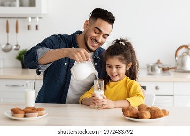 Snack Time. Happy Arab Dad And Daughter Eating Pastry And Drinking Milk In Kitchen, Cheerful Middle Eastern Daddy And Child Having Fun Together, Caring Father Pouring Calcium Drink From Jug To Glass