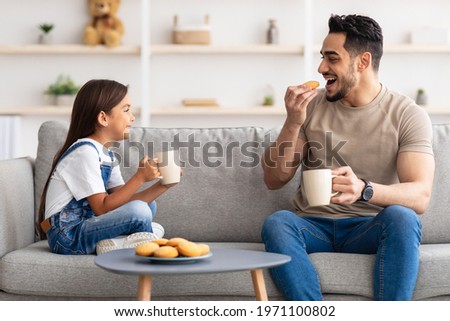 Snack Time. Portrait of cheerful little girl and man holding cups, drinking coffee and tea, eating cookies and looking at each other, dad and daughter talking sitting on the couch in living room