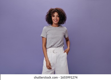 Snapshot Of Curly Woman In White Shorts Against Purple Background. Charming Brunette Girl In Grey Tee Smiling On Isolated Backdrop